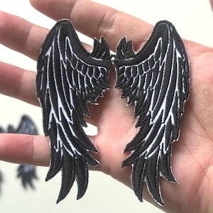 Puede incluir: Dos parches de alas bordadas en negro con detalles blancos. Los parches tienen forma de alas de ángel, con cada pluma cosida individualmente. Las alas se muestran sobre un fondo neutro.