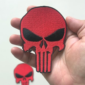 Puede incluir: Parche bordado rojo y negro con un diseño de calavera. El parche presenta una calavera roja con cuencas de ojos negras y un contorno negro. Una versión más pequeña del parche es visible en el fondo.