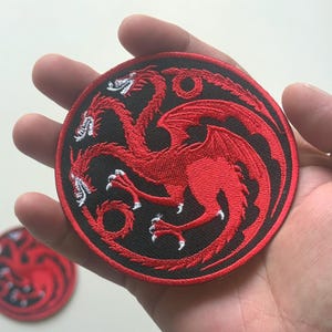 May include: Round, embroidered patch featuring a red three-headed dragon design on a black background. The patch is held in a hand, with a second patch visible in the background. The dragon has detailed wings and claws.
