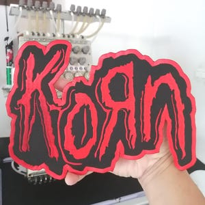 May include: A fabric patch featuring the band name Korn embroidered in a jagged, stylized red font against a solid black background. The lettering has a distinct, distressed aesthetic with a thick red border outlining the entire shape.