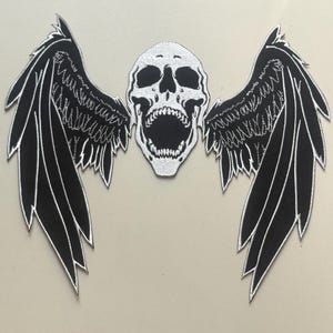 Pode incluir: Um patch preto e branco com uma caveira com asas de penas. A caveira é branca com detalhes pretos, e as asas são pretas com acabamento branco. Ideal para roupas ou acessórios.