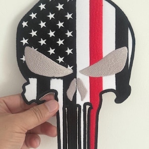 May include: Embroidered patch in the shape of a skull, featuring the American flag design on the left side with white stars on a black background. The right side has white and red stripes. The skull's eyes are gray.
