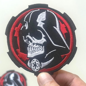 May include: Embroidered patch featuring a black and white depiction of Darth Vader's helmet within a red and black circular frame. The patch includes a symbol at the bottom and is likely for clothing or accessories.