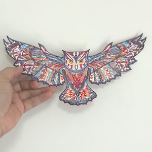 Op de afbeelding: Een kleurrijke geborduurde uil patch met uitgestrekte vleugels. De patch heeft ingewikkelde patronen in tinten blauw, oranje, rood en paars. De uil heeft gele ogen en een rode snavel. De patch wordt tegen een witte achtergrond gehouden.