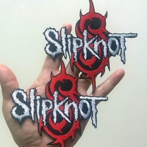 Pode incluir: Dois patches bordados com o nome da banda "Slipknot" em letras brancas estilizadas. Os patches têm um design vermelho e preto em forma de chama em torno do texto. Os patches são segurados em uma mão.
