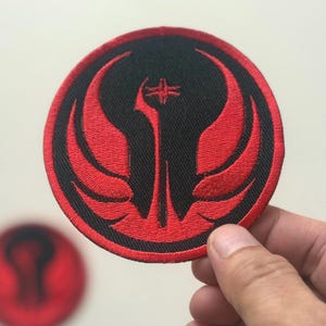 May include: A round, embroidered patch featuring a black background with a red emblem. The emblem has a central vertical element with a cross-like design at the top, flanked by stylized wings. The patch is held by a hand.
