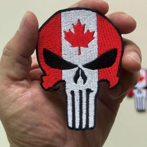 May include: A round patch featuring a skull design with the Canadian flag incorporated. The top half of the skull is the Canadian flag, with red and white stripes and a red maple leaf. The bottom half is a white skull with black eye sockets.