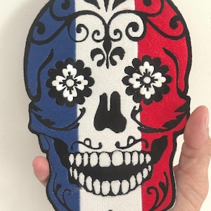 May include: Embroidered skull patch with a design inspired by the French flag. The skull features blue, white, and red vertical stripes, with black floral accents and a detailed black outline. The eyes are represented by white flowers with black centers.
