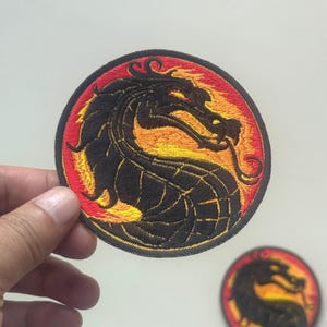 Può includere: Toppa ricamata con un disegno di drago nero su uno sfondo circolare rosso e giallo. Il drago è dettagliato con cuciture intricate. È visibile anche una versione più piccola della toppa.