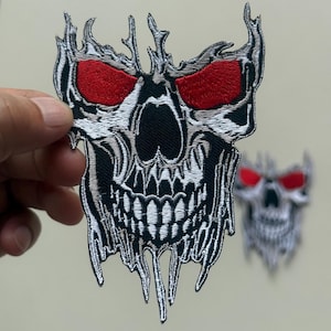 May include: Embroidered skull patch with red eyes, black nose, and white teeth. The skull has a detailed design with a dripping effect. A second, blurred patch is visible in the background. This patch is suitable for jackets, bags, or other apparel.