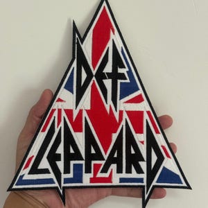 以下が含まれることがあります： 三角形のDef Leppardパッチ。パッチは、赤、白、青のユニオンジャックのデザインを背景に、黒の様式化された文字でバンド名を表示しています。パッチには白い縁があります。