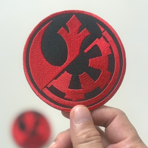 Peut inclure: Un écusson rond brodé avec un symbole rouge sur fond noir. Il est tenu par une main sur fond blanc. Un autre écusson flou est visible en arrière-plan. Le motif est un emblème stylisé.