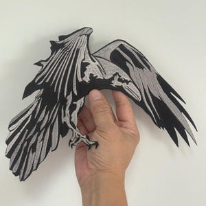 May include: Embroidered raven patch in black and grey, depicting a bird with spread wings. The patch has detailed stitching, giving it a textured look. The patch is held against a white backdrop.