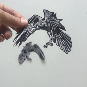 May include: Embroidered raven patches in black and grey. The larger patch shows the raven in flight with outstretched wings. The smaller patch is a raven in flight. The patches are held by a hand against a white background.
