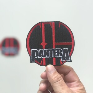 May include: A black and red embroidered patch with the word "PANTERA" in white lettering. The patch has a circular design with red accents and a stylized "T" in the center. A second, blurred patch is visible in the background.
