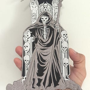May include: Embroidered patch featuring the Grim Reaper in a hooded cloak, holding a scythe. The design includes a skeletal figure seated on a throne with skull accents. The patch is primarily black and gray, with detailed stitching.