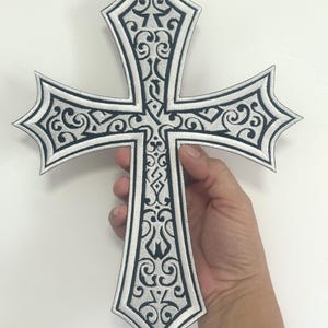 Op de afbeelding: Een groot, wit kruis met een zwart, sierlijk, geborduurd ontwerp. Het kruis heeft puntige randen en wordt tegen een witte achtergrond gehouden. Het ontwerp heeft wervelende patronen en een gedetailleerde rand.