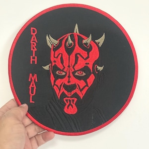 May include: Embroidered patch featuring Darth Maul from Star Wars. The patch is circular with a black background and a red border. The image of Darth Maul is in red, with his face and horns prominently displayed. The text "Darth Maul" is vertically embroidered.