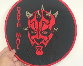 Può includere: Toppa rotonda ricamata raffigurante Darth Maul di Star Wars. La toppa ha uno sfondo nero e un bordo rosso. L'immagine di Darth Maul è rossa, con il suo volto e le sue corna ben visibili. Il testo "Darth Maul" è ricamato verticalmente.