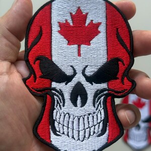 May include: Embroidered patch depicting a skull design incorporating the Canadian flag. The skull is white with black eyes and a detailed jawline. The flag's red and white colours are prominent, with a red maple leaf in the centre.