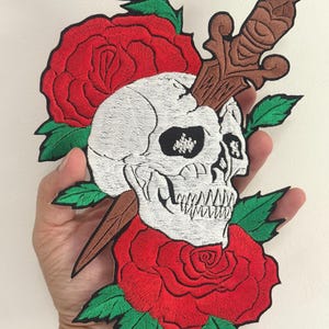 May include: Embroidered patch featuring a white skull pierced by a brown dagger, surrounded by two red roses with green leaves. The patch has a detailed, textured design, suitable for jackets, bags, or other fabric items.