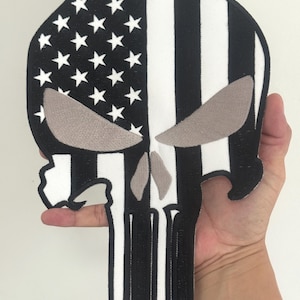 May include: A black and white skull patch with a design inspired by the American flag. The patch features white stars on a black background, white and black stripes, and gray eye shapes. The skull's jawline is visible.