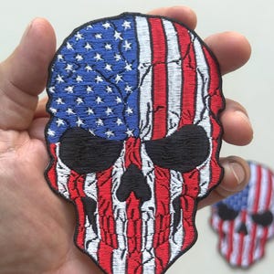 May include: Embroidered skull patch featuring the American flag design. The top half of the skull is blue with white stars, and the bottom half has red and white stripes. The patch is held in a hand, with a second patch visible in the background.