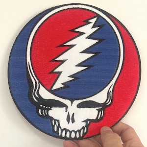 Może przedstawiać: Okrągła naszywka z czaszką i błyskawicą. Projekt jest w kolorach czerwonym, białym i niebieskim. Czaszka ma białą twarz i czarne kontury. Naszywka ma średnicę około 13 cm.