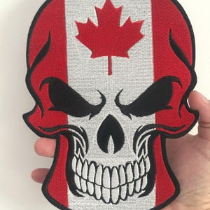 May include: Embroidered skull patch featuring the Canadian flag design. The skull has a white face with black eyes and a menacing grin. The top of the skull is red and white, with a red maple leaf in the center.