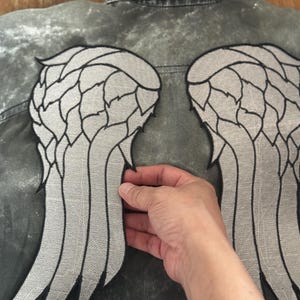 Puede incluir: Chaqueta vaquera gris con grandes parches bordados de alas de ángel en la espalda. Las alas son de color gris claro con contornos negros, creando un contraste llamativo con el color descolorido de la chaqueta.