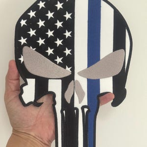 Può includere: Una toppa a forma di teschio nera, bianca e blu con il design della bandiera americana sul lato sinistro e una striscia blu sul lato destro. Il teschio ha dettagli grigi per gli occhi e il naso. La toppa è tenuta in mano.