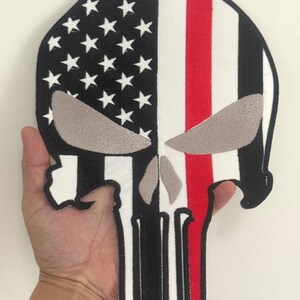 May include: A skull-shaped patch with an American flag design. The left side features a black background with white stars, while the right side has red and white stripes. The eyes are gray, and the outline is black.