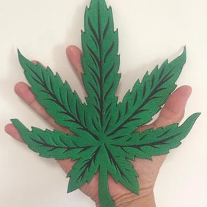 Op de afbeelding: Een grote, groene cannabisblad-vormige patch met zwarte aderdetails. De patch wordt in een hand gehouden tegen een witte achtergrond. Het blad heeft een getextureerd uiterlijk.