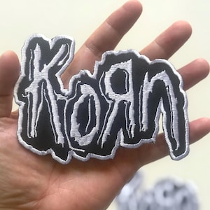 Puede incluir: Parche bordado negro con la palabra "KORN" en una fuente estilizada, blanca y goteante. El parche tiene un fondo negro y un borde blanco. El parche se sostiene en una mano.