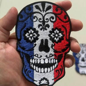 May include: Embroidered skull patch featuring the French flag colors: blue, white, and red. The skull design includes floral accents and a detailed tooth structure. This patch is suitable for jackets, bags, or other fabric items.