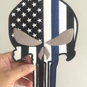 May include: A skull-shaped patch featuring the American flag design on the left side with white stars on a black background. The right side has a blue stripe, and the skull's eyes are embroidered in gray. The patch is held by a hand.
