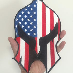 Op de afbeelding: Een patriottische patch in de vorm van een gestileerde helm. Het ontwerp bevat de Amerikaanse vlag, met blauwe sterren en rode en witte strepen. Zwarte accenten definiëren de kenmerken van de helm.
