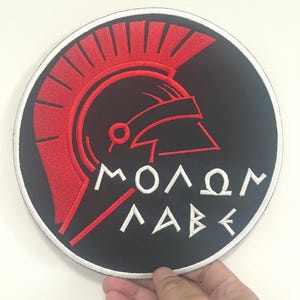 Op de afbeelding: Ronde zwarte patch met een rood Spartaans helm ontwerp en de Griekse zin "ΜΟΛΩΝ ΛΑΒΕ" geborduurd in wit. De patch heeft een witte rand.