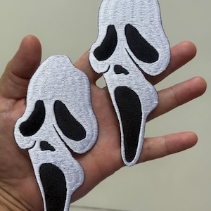 May include: Two white and black Scream mask patches. The patches feature a white embroidered design with black eye and mouth details. The patches are likely for clothing or accessories.