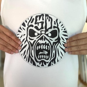 Peut inclure: Un débardeur blanc avec un grand écusson circulaire. L'écusson présente un graphisme noir et blanc d'un visage menaçant avec des dents acérées et une expression menaçante. Le débardeur est uni.