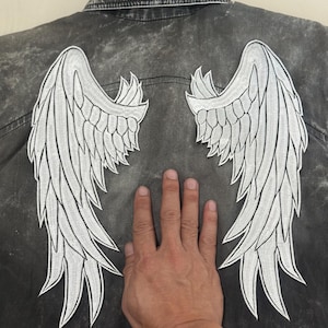 Puede incluir: Chaqueta vaquera negra descolorida y desgastada. La parte trasera de la chaqueta presenta grandes alas de ángel blancas bordadas. Las alas están detalladas con patrones de plumas individuales.