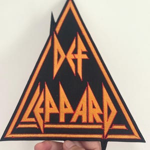 以下が含まれることがあります： 黒とオレンジ色のDef Leppardパッチ、三角形の形。バンド名とロゴは、黒い背景にオレンジ色の糸で刺繍されています。パッチは、上部と下部が尖っています。