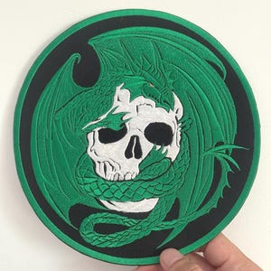 May include: Embroidered patch featuring a white skull encircled by a green dragon on a black background. The dragon's wings are spread, and its body wraps around the skull. The patch is circular and has a black border.