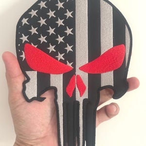 May include: Embroidered patch in the shape of a skull with an American flag design. The left side features white stars on a black background, while the right side has black and gray stripes. The skull's eyes and nose are red.