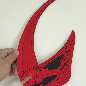 May include: A red, stylized, horned mask with black accents. The mask has a curved, elongated horn and a detailed, skull-like design. The mask is held against a white background.