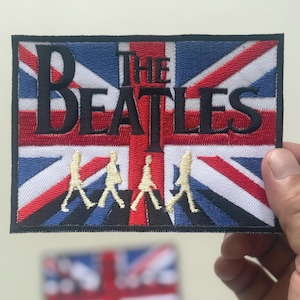 Könnte beinhalten: Gestickter Aufnäher mit dem Design der Union-Jack-Flagge und dem Text "THE BEATLES" in schwarzer Schrift. Darunter befinden sich vier gelbe Silhouetten der Bandmitglieder beim Gehen. Der Aufnäher ist rechteckig mit schwarzem Rand.