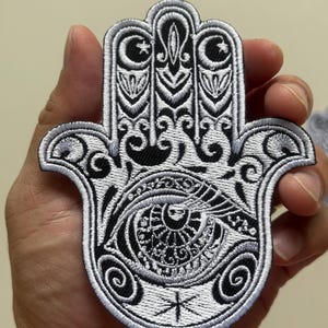 Könnte beinhalten: Gestickter schwarz-weißer Hamsa-Hand-Aufnäher. Der Aufnäher zeigt ein Augendesign in der Mitte mit komplizierten Details und einem dekorativen Rand. Das Design umfasst Halbmonde, Sterne und wirbelnde Muster.