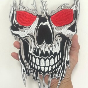 May include: Embroidered skull patch with red eyes, black mouth and nose, and white and gray detailing. The patch has a jagged, flame-like border. The patch is held in a hand.
