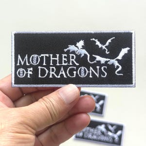 May include: Black rectangular patch with white embroidered text and dragon silhouettes. The text reads "MOTHER OF DRAGONS 2". The patch has a white border and is held by a hand.