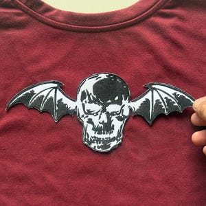 Peut inclure: Un t-shirt bordeaux avec un écusson de crâne noir et blanc avec des ailes de chauve-souris. L'écusson est centré et présente un motif détaillé. Le t-shirt est fait d'une matière douce.